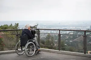 Disability Prints: Senior man in wheelchair looking at city through coin operated binoculars from viewing balcony Observatory Griffith
