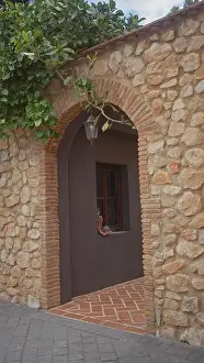 Gaze Prints: A senior woman peeks curiously through a rustic stone archway in a charming mediterranean alley