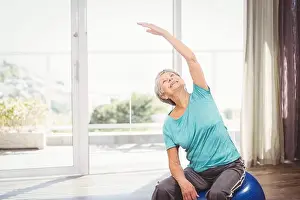 Flexibility Prints: Senior woman is stretching on exercise ball by sliding glass door opening to terrace with plants