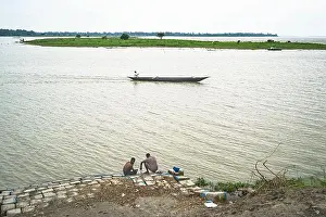 Slow Living Prints: Serene Riverside Life in Rural Bangladesh – Two Men Resting by the Jamuna River as a Traditional Boat Passes by