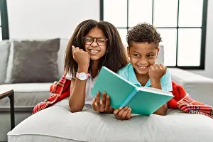 Sibling Prints: Two siblings lying on the sofa reading a book very happy and excited doing winner gesture with arms raised