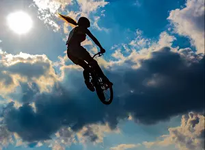 High Jump Prints: Silhouette woman of a cyclist flying against a blue sky with clouds and sun