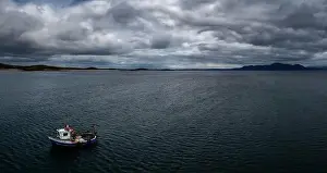 County Mayo Prints: Single fishing boat in a deep blue ocean under a cloudy sky with coastal mountains in the background