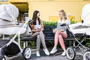 Disposable Prints: smiling mothers sitting on bench with coffee to go near baby strollers
