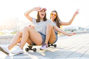 Female Friends Prints: Two smiling young girls having fun while riding on a skateboard at the park