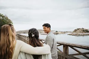 Female Friends Prints: Smiling young man with female friends walking on bridge near lake