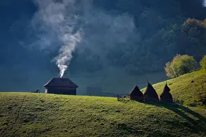 Smoke raising from the chimney of a lonely old wood house near three haystacks on a mountain hill