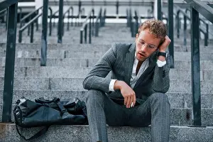 Nervous Prints: Smoking cigarette. Young businessman in grey formal wear is outdoors in the city