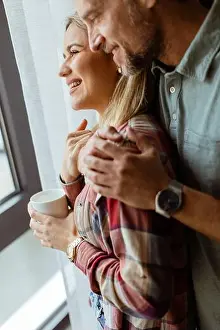 Contentment Prints: In a softly lit room, a man lovingly embraces a woman from behind as they both gaze out the window