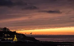Isle Of Wight Prints: a streak of bright orange light cutting across a sun set dark sky on the coast with an outside pine Christmas tree