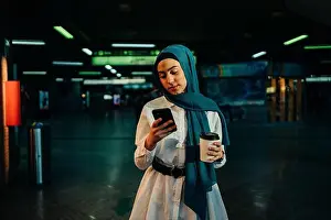 Headwear Prints: Stylish Muslim female in hijab standing with takeaway coffee in subway and surfing Internet on mobile phone