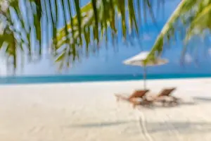 Summer exotic sandy beach with blur palms and sea on background. Blurry beach landscape, chairs and umbrella under palm leaves