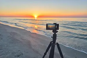 Sunrise over the sea coast. Fireball of the sun above the horizon in a colorful orange sky. Smartphone camera on a tripod in the foreground