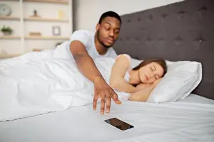 Betrayal Prints: Suspicious black husband checking his sleeping wife's cellphone in bed at home