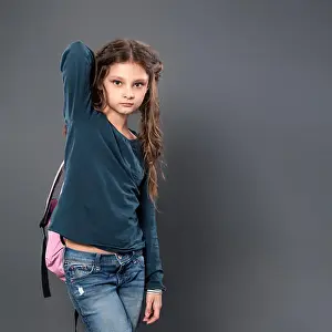 Thinking pupil kid gilr posing in style clothing and holding backpack on grey studio background. Closeup