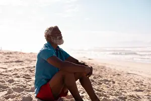 Pensive Prints: Thoughtful bearded african american senior man with gray hair sitting on sandy beach against sky