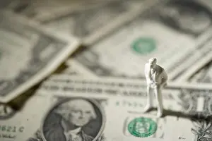 A tiny white figurine of a man in a business suit stands on a bed of US dollar bills