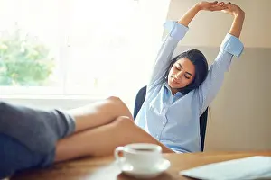 Exhausted Prints: Tired young woman with feet on desk and stretching arms upward at home office
