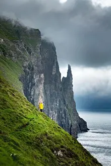 Tourist in yellow jacket looks at Witches Finger cliffs from Trollkonufingur viewpoint. Vagar island, Faroe Islands, Denmark