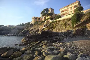 Holiday Maker Prints: Town buildings on a cliff facing the sea at sunset seen from a cove