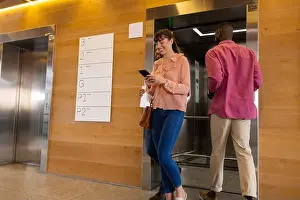 Texting Prints: Using smartphone, woman walking past man entering elevator in office building