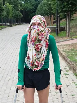 Summer Trees Prints: Vertical shot of a gender-fluid person wearing a floral niqab and walking on crutches in a park
