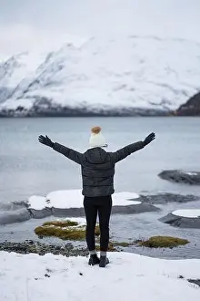 Back view of anonymous Woman in warm down jacket and hat with outstretched arms standing on beach against white