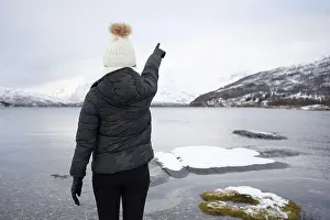 Unrecognizable Prints: Back view of anonymous Woman in warm down jacket and hat pointing with finger while standing on beach against white
