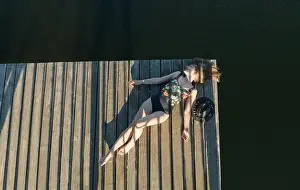 Faceless Prints: Top view of beautiful Woman in black swimsuit and hat lying down on wooden pier of lake on clear blue sky