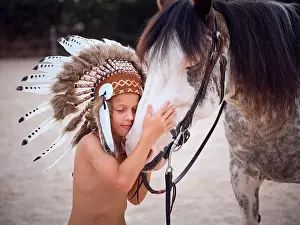Headwear Prints: Side view of cute child with closed eyes wearing traditional Indian headwear with feathers caressing muzzle of gray