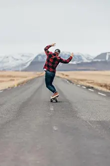 Unrecognizable Prints: Back view of hipster guy riding skateboard on paved long road with snowy mountains on background in Iceland