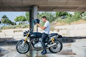 Assured Prints: Side view of male biker sitting on motorcycle parked under bridge and putting on protective helmet
