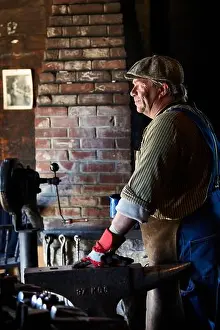 Craft Prints: Side view of middle aged man in apron and glove standing near anvil and looking away against brick wall in smithy