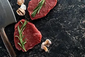 Rosemary Prints: top view of raw beef steaks with rosemary twigs and garlic cloves near sharp knife on black marble surface