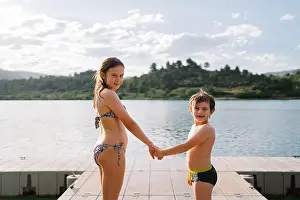 Sibling Prints: Side view of smiling brother and sister in swimwear standing together holding hands on quay near lake