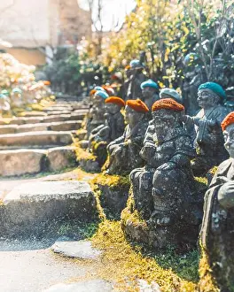 Spirituality Prints: View of stone Jizo statues wearing colorful knitted caps lined along a mossy stone staircase in a temple garden
