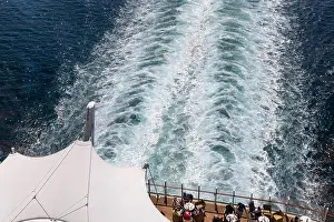 Wake Prints: View from upper deck showing the foaming wake trailing behind a cruise ship as passengers relax at tables on the aft