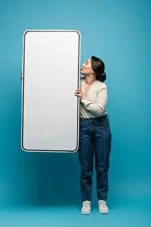 Cardigan Prints: Side view of young asian woman pouting lips and holding big smartphone model on blue background