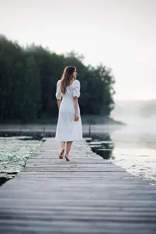Calming Prints: Back view of young woman in white dress standing alone on footbridge and staring at lake