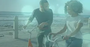Walking couple holding bicycles on seaside promenade, with ocean waves and metal railing