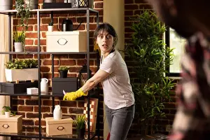 Cleanliness Prints: White woman wearing yellow rubber gloves wipes dust from shelves while listening to boyfriend