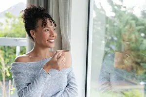 Contentment Prints: Woman in her 30s standing by window at home holding ceramic mug wearing off shoulder sweater