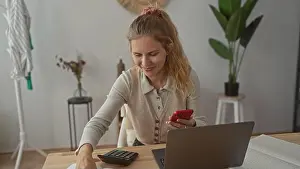 Stool Prints: Woman balances phone while calculating budgets in a bright office with a blonde young home setup featuring laptop