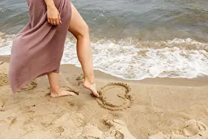 woman in beige dress draws with her foot heart on the sand by sea on summer day. outdoor closeup