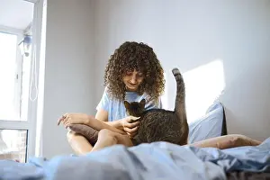 Satisfaction Prints: A woman with curly hair sits on a bed while gently petting a cat