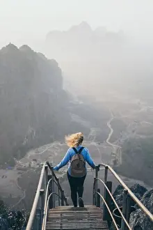 Woman in denim shirt standing on mountain peak with stairs going down during sunrise foggy morning in Hpa-An, Myanmar