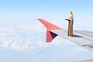 Woman in dress and hat standing on airplane wing and working on laptop