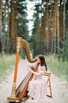 Tune Prints: Woman harpist sits at forest road and plays harp in beautiful dress against a background of pines