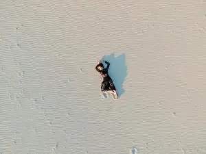 Woman in lace black dress lies in desert and casts a shadow on white sand. View from drone directly above