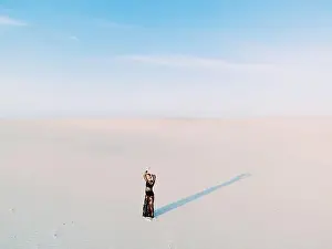 Woman in lace black dress stands in desert and casts a shadow on white sand. View from drone